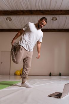 Athletic man in white t-shirt and sweatpants stetching his legs, warming up before training, side view close up portrait. standing at hall in his apartment workout program he found online.