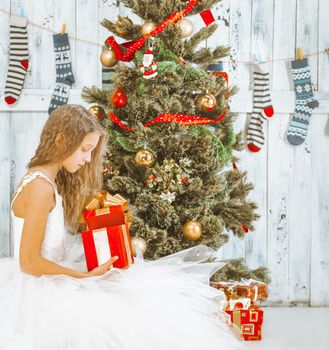 Teenage Girl with Long Hair in a White Tutu Dress Sits under a Christmas Tree and Unpacks her Present. Christmas Morning. High quality photo