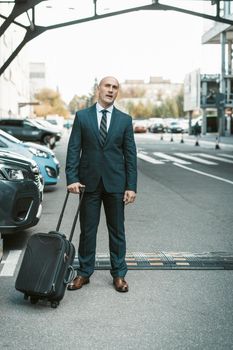 Waiting for his driver or cab handsome businessman stands on parking lot carrying suitcase. Businessman in suit and suitcase on business trip. Man standing outdoors with luggage.