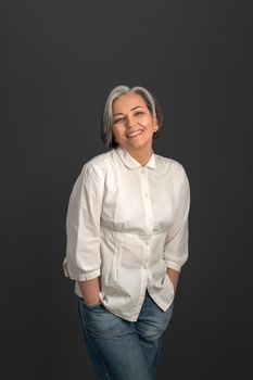 Senior grey haired charming woman looking happy wearing white shirt and denim jeans on camera isolated on grey background. Portrait of mature woman in casuals looking happy on camera.