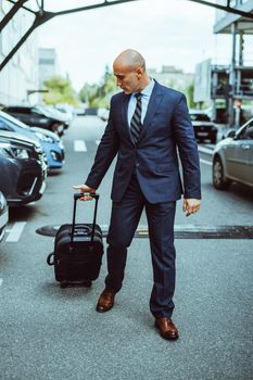 Bald businessman walking on the airport parking lot with suitcase and parked cars. Businessman in suit and suitcase on business trip. Man walking outdoors with luggage determined and confident.