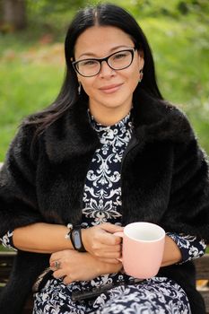 Close up charming asian woman in fur coat and beautiful dress sits at park on old rusty bench with a pinkish cup of coffee or tea. Resting woman in sunny day outdoors.