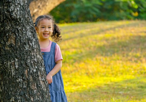 Asian little girl stay behind the big tree and smile with morning light in garden.