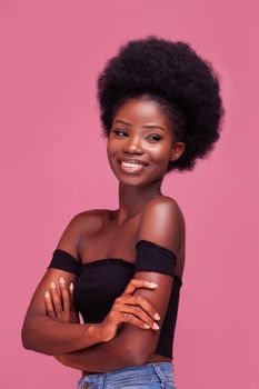 Happy charming African American girl with amazing afro hair standing smiling with arms folded in black top and denim jeans isolated on pink background.