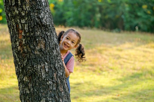 Little Asian girl play peekaboo by hiding back of tree and smile, she play in green garden with morning light.