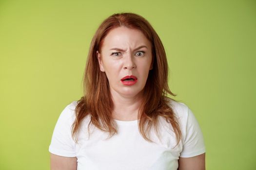 Confused shocked gasping middle-aged redhead woman cringe frustrated puzzled open mouth speechless freak out strange shocking scene stand green background perplexed disappointed. Copy space