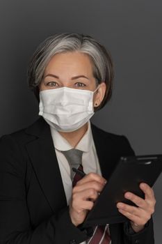 Caucasian grey haired businesswoman in protective mask work using digital tablet gadget looking at camera isolated on grey background. Studio shot.