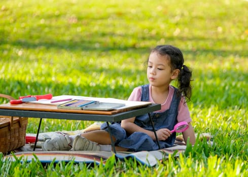 Cute Asian little girl hold magnifier glass and sit near table with colored pencils and toys in the garden with morning light.