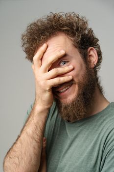 Shy handsome young man covered his face with hand pry with his eye, curly hair in olive t-shirt isolated on white background. Portrait of smiling young man.