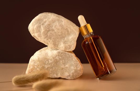 Transparent glass cosmetic dropper standing near stones and dry flowers on dark red and trendy beige background. Beauty composition. Concept of natural and original skincare products presentation.