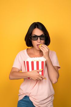 Young brunette woman with bucket of popcorn in stereo glasses. Cinema concept. Isolated on yellow background.