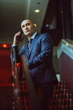 Middle aged bald businessman stopped on the stairs holding smartphone using mobile phone in the office. Male boss caucasian man is giving instructions to his employees with concentrated look away.