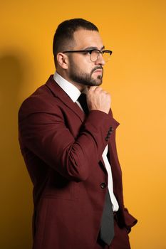 Handsome young man eye glasses wearing burgundy suit looking seriously away with hand folded touching chin standing sideways isolated on yellow background.