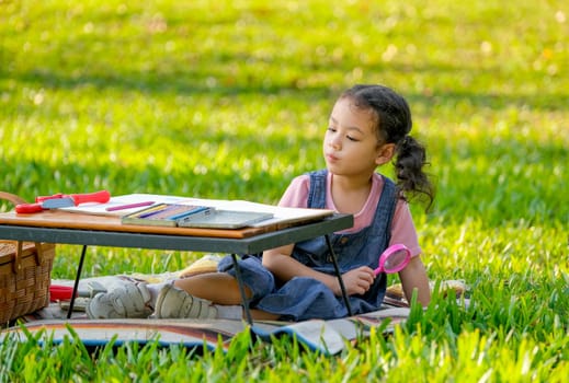 Portrait of Asian little girl hold magnifier glass and sit near table with colored pencils.