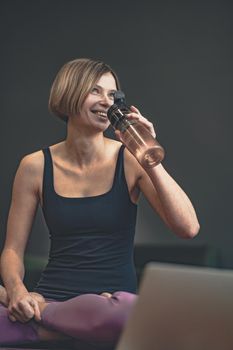 Smiling Woman Drinks Water from a Bottle and Sits on Floor with Crossed Legs in Sportswear. Replenish Water Balance, Monitor the Water Level in the Body, Saturate the Body with Water.