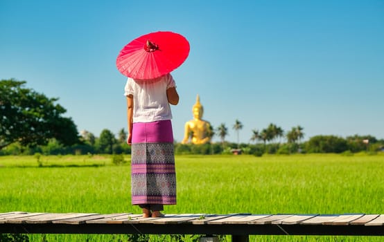 Woman with traditional skirt and red umbrella stand on wood bridge in front of green rice field and she look to Buddha statue with day light and blue sky.