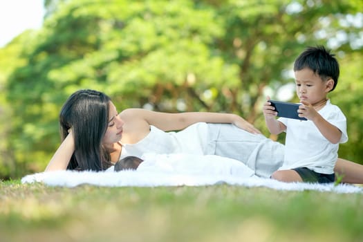 Young Asian mother relax by lie on white carpet near her little baby and her son who enjoy with mobile phone and internet.