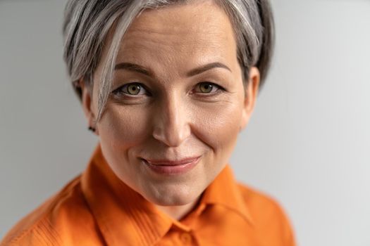 Close up portrait of a happy mature grey haired woman wearing orange shirt looking at camera. Pretty mid aged grey haired woman in orange shirt isolated on grey background.