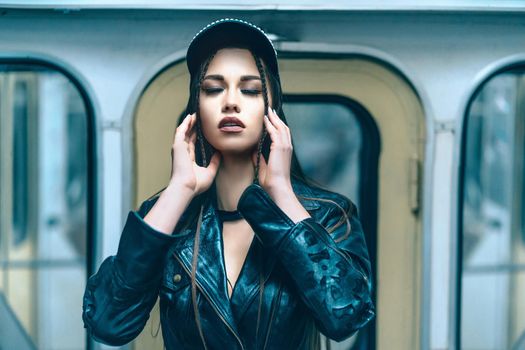 Attractive Glamorous Model Female with Close Eyes Posing Inside the Metro Train. Cute Young Woman in Leather Jacket and Modern Cap Inside the Subway Car. Close-up Portrait. Windows Background. High quality photo