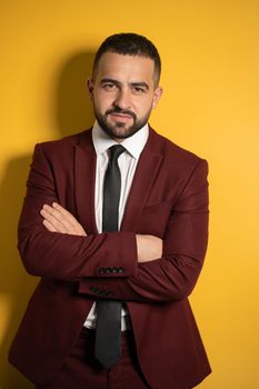 Half-length portrait of handsome man in burgundy color suit looking at camera with hands folded isolated on yellow background. Vertical photo.