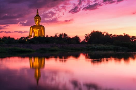 Reflection of big buddha statue in the river with purple sunset light and some clouds in the sky.