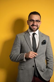 Handsome bearded smiling elegant man wearing eye glasses in formal wear with one hand lifted standing isolated on yellow background.