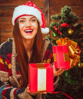 Caucasian Brunette Lady with a Wide Smile in a Casual Sweater and a Santa Hat Holds an Open Red Gift Box in her Hands Next to the Christmas Tree. Christmas Gifts, Christmas Mood. Wood Background. High quality photo