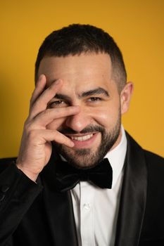 Eye cover one hand handsome bearded man in tuxedo hide peeps out on closed eyes. Handsome young smiling caucasian man isolated on yellow background. Vertical photo.