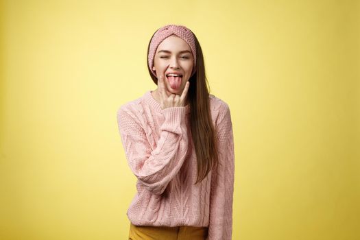 Silly timid-looking girl having fire rebellious heart inside showing rock-n-roll gesture sticking out tongue sassy winking at camera, smiling having fun at heavy-metal concert over yellow background.