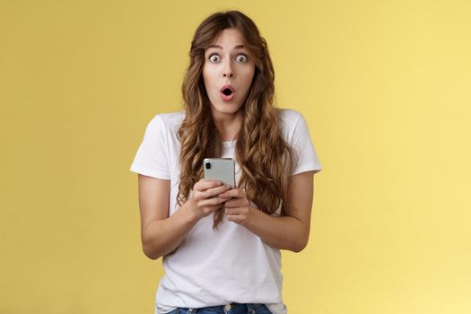 Shocked impressed speechless girl say wow curly hairstyle stare camera astonished hold smartphone reacting stunning perfect news receive awesome message stand yellow background. Copy space