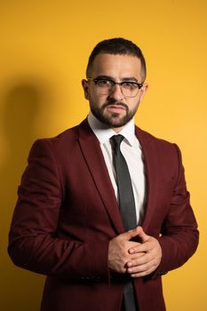 Half-length portrait of handsome man standing half turn in burgundy color suit and eye glasses looking at camera with hands folded isolated on yellow background. Vertical photo.