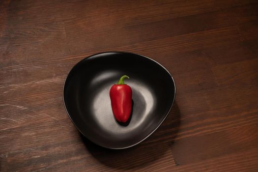 Organic mini red bell pepper laying lonely in a deep black bawl and ready to eat isolated on wooden background.