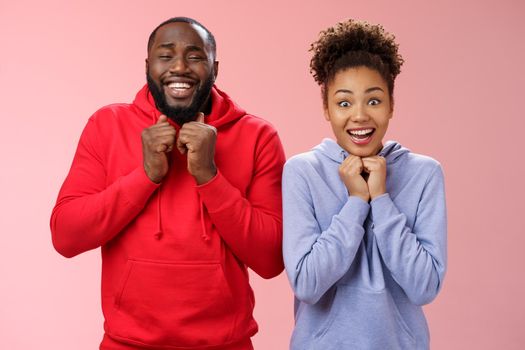 Excited happy girlfriend boyfriend smiling broadly nervously clenching fists pressed chest looking camera thrilled anticipate good news trying luck playing lottery together, standing pink background.