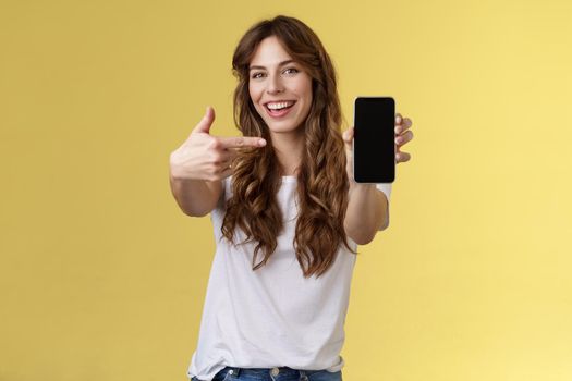 Cheerful sassy good-looking sociable girl curly long hair extend arm holding smartphone pointing index finger mobile phone screen smiling broadly recommend cool app blogger promote social page. Lifestyle.