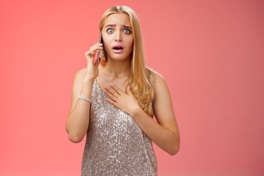 Lifestyle. Concerned worried woman blond hairstyle in silver stylish glittering dress hold hand heart widen eyes afraid gasping speechless listen terrifying news talking smartphone express empathy worry.