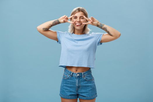 Enjoy life and dream. Portrait of joyful good-looking carefree european female student with blond short haircut winking sticking out tongue entertained and showing peace signs over eyes near blue wall.
