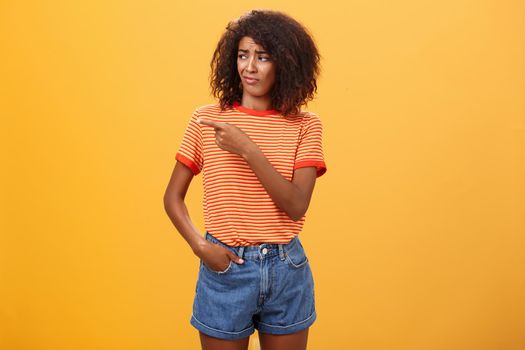 Girl looking at doubtful unimpressive perfomance of model. Portrait of displeased confused good-looking dark-skinned female with afro hairstyle looking and pointing left with scorn and indifference.