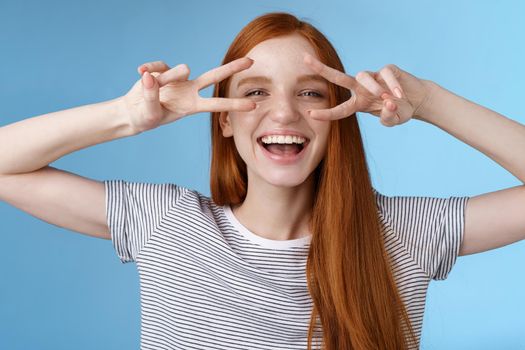 Happy cheerful carefree caucasian redhead girl ginger long hair having fun express happiness joy friendly attitude show peace victory gesture disco signs eye laughing, standing blue background.