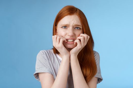Worried uncomfortable scared young panicking redhead young girl feeling pressure distressed frowning squinting frightened biting fingernails trembling fear, standing blue background.