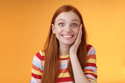 Cheerful glad excited attractive redhead girl blushing surprised feeling happy touch cheek pleased receive good news standing joyful thrilled get awesome chance, posing orange background.