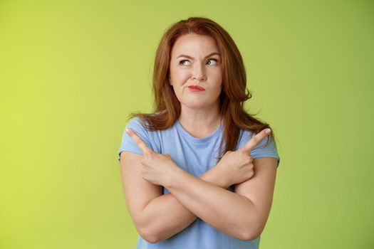 Time make right choice. Perplexed hesitant redhead middle-aged female customer deciding between few variants cross arms body pointing sideways look left smirking unsure doubting choosing product.