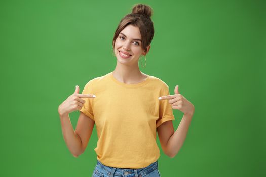 Calm and polite friendly-looking skillful young female coworker in yellow t-shirt pointing at herself self-assured tilting head and smiling at camera wanting participate in event over green background. Lifestyle.