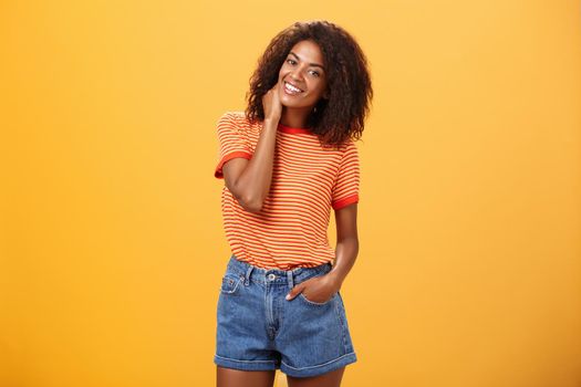 Stylish African American slim woman with curly medium hair holding hand in denim shorts tilting head, rubbing neck while smiling friendly feeling uncomfortable or shy in new company over orange wall.