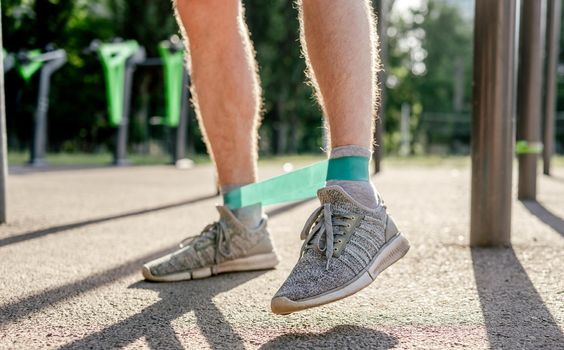 Closeup side view of legs feets of man guy training with elastic rubber band at the stadium outdoors. Athlete adult male person during active workout with additional sport equipmant