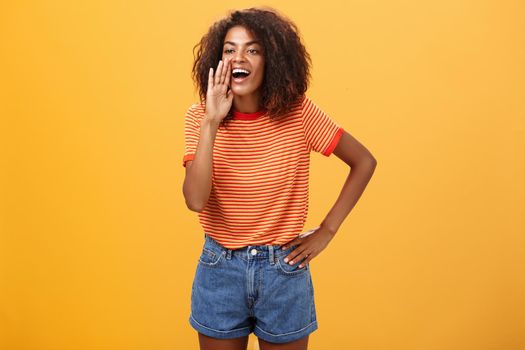 Girl calling sister come to phone yelling out loud holding palm near opened mouth to be scream louder so friend can hear holding arm on waist gazing left with carefree pleased look over orange wall. Lifestyle.