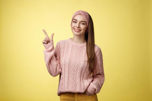 Girl picked staff, made decision pointing up at product smiling pleased. Studio shot of young student in knitted sweater, grinning indicating upper left corner enthusiastic, giving recommendation.