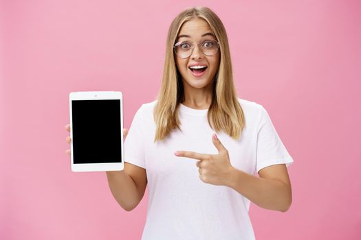 Girl suggests buy digital tablet for university and forget paper books. Excited happy and delighted young female student in white t-shirt and glasses pointing at device screen recommending gadget. Technology, emotions, advertising and lifestyle concept