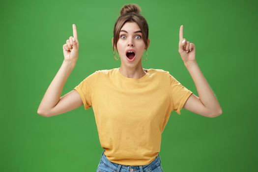 Waist-up shot of interested and surprised amazed good-looking female in yellow t-shirt with tattoo dropping jaw impressed and astonished raising hands pointing up questioned against green background. Copy space