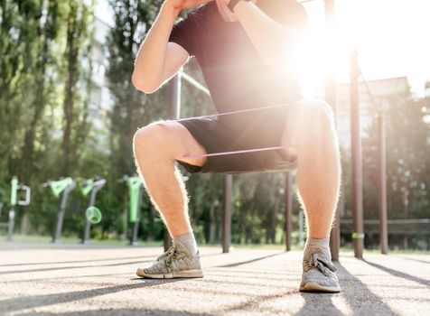 Strong sportive man guy doing squats with elastic rubber band at the stadium outdoors. Adult male person during workout for legs with additional sport equipmant. Sportsman exercising outside