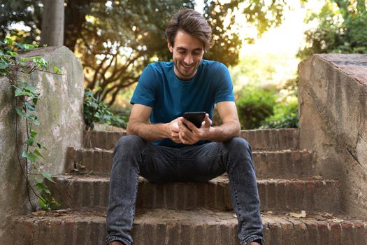 Happy young caucasian man sitting on stairs using mobile phone browsing social media in nature. Copy space. Lifestyle and social media concept.